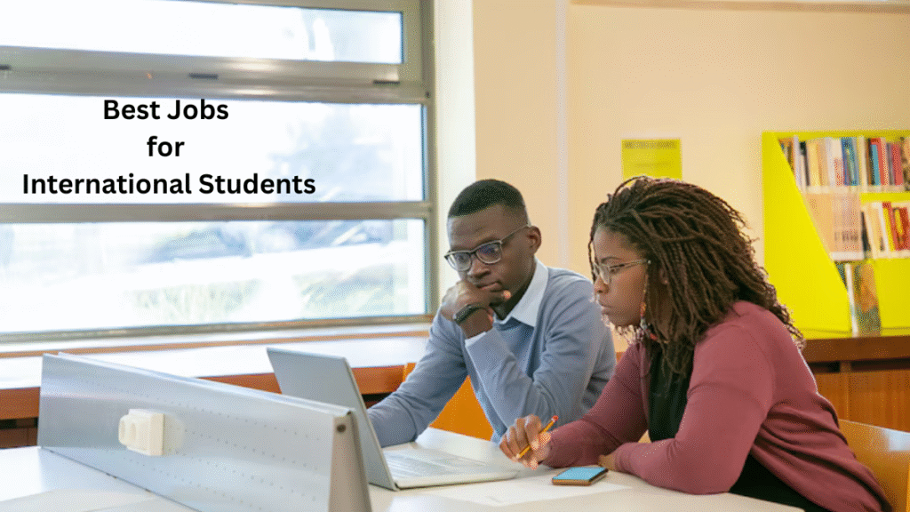Two students working together at a desk in a library, using a laptop and taking notes, with the text “Best Jobs for International Students” displayed on the image.