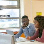 Two students working together at a desk in a library, using a laptop and taking notes, with the text “Best Jobs for International Students” displayed on the image.